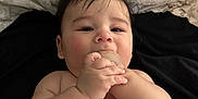 Jason is registered to the contest to win money with this photo: baby, infant, chewing, hand, bare_skin, lying_down, dark_hair, curious_eyes, chubby_cheeks, patterned_background, black_surface, cute, child, one_person, portrait, indoors, skin, young_child, baby_hand, baby_face