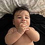 baby, infant, chewing, hand, bare_skin, lying_down, dark_hair, curious_eyes, chubby_cheeks, patterned_background, black_surface, cute, child, one_person, portrait, indoors, skin, young_child, baby_hand, baby_face