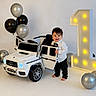 toddler, child, toy_car, mercedes, balloons, birthday, number_one, white_background, celebration, smiling, barefoot, formal_clothing, bow_tie, party, indoor, happy, curly_hair, black_pants, white_shirt, lighting