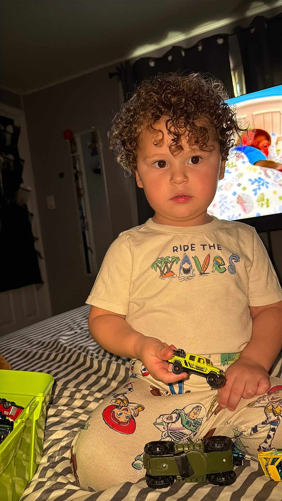 Jordao joined the competition — help win amazing prizes! child, toddler, curly_hair, toy_car, bed, striped_bedspread, pajamas, tshirt, indoor, bedroom, television, screen_image, sitting, playtime, toys_box, blanket, closeup, flash_lighting, young_child, facial_expression
