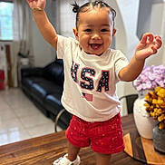 ángela is registered to the contest to win money with this photo: toddler, child, smiling, standing, table, usa_tshirt, red_shorts, white_shoes, indoor, living_room, flowers, vase, wooden_table, happy, person, cute, hair_pigtails, portrait, home, bright