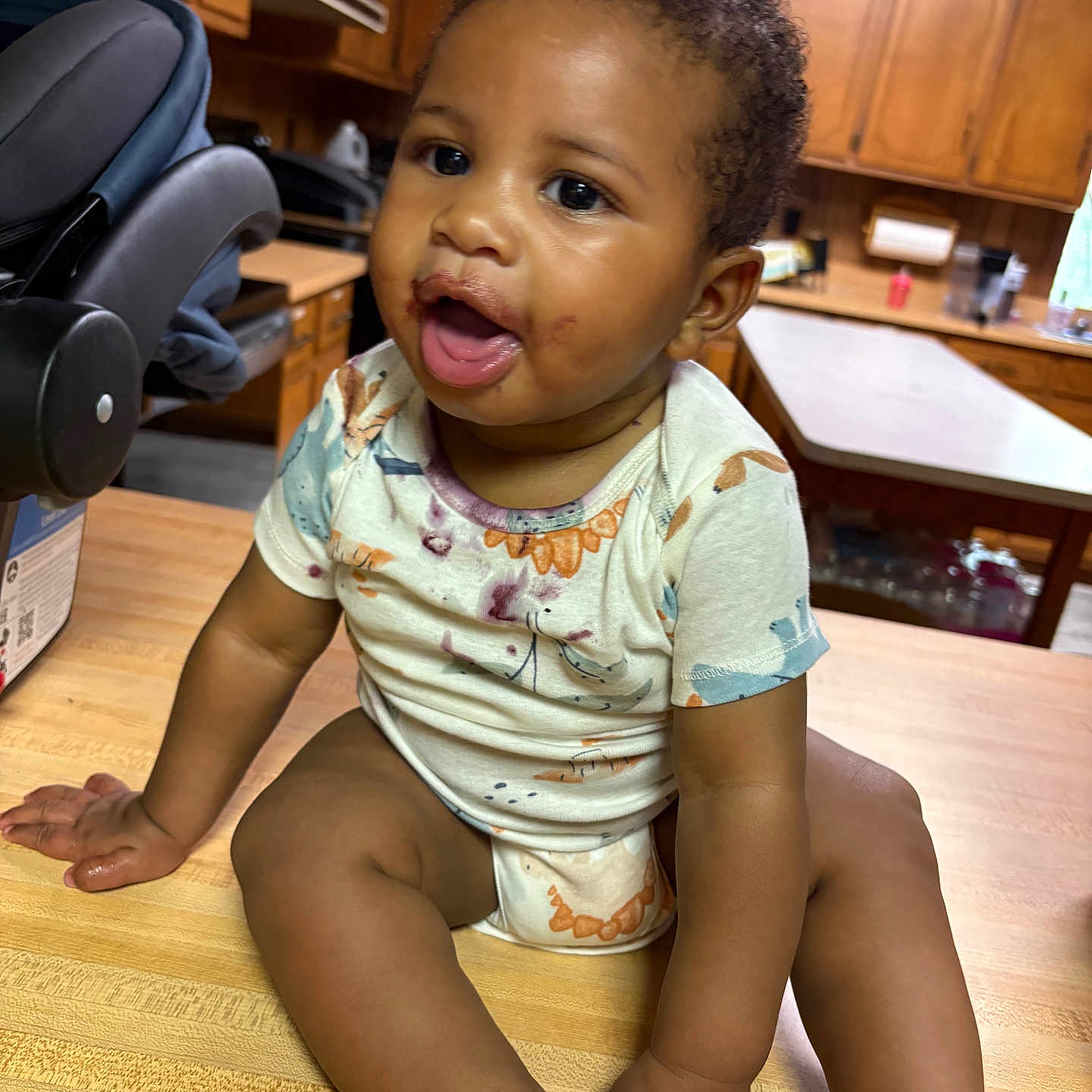 Cj is registered to the contest to win money with this photo: baby, infant, child, kitchen, counter, wooden_surface, messy_face, curly_hair, short_sleeve_shirt, sitting, indoors, happy, cute, person, cabinet, appliance, table, home, curious, smiling