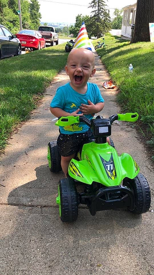 Desmond is registered to the contest to win money with this photo: asphalt, auto_part, automotive_tire, automotive_wheel_system, car, fun, grass, lawn, motor_vehicle, person, plant, recreation, riding_toy, smile, tire, toddler, toy, tread, tree, vehicle
