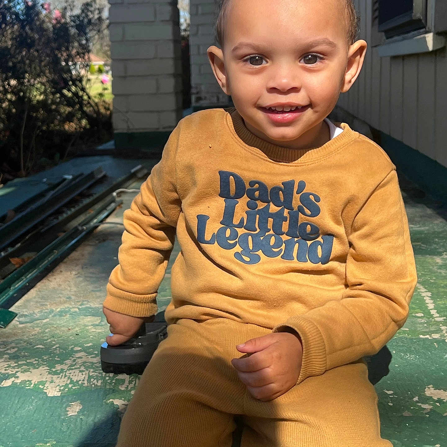 Marley is registered to the contest to win money with this photo: child, toddler, smiling, outdoor, porch, sweatshirt, pants, yellow_clothing, sunlight, happy, person, face, short_hair, holding_object, concrete, green_floor, daylight, casual, cute, young_child