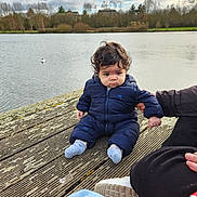 Lucas participe au concours pour gagner de l'argent avec cette photo : baby, toddler, child, dock, wooden_planks, lake, water, jacket, snowsuit, socks, adult_hand, shoe, cloudy_sky, trees, nature, outdoors, portrait, curly_hair, seagull, expression