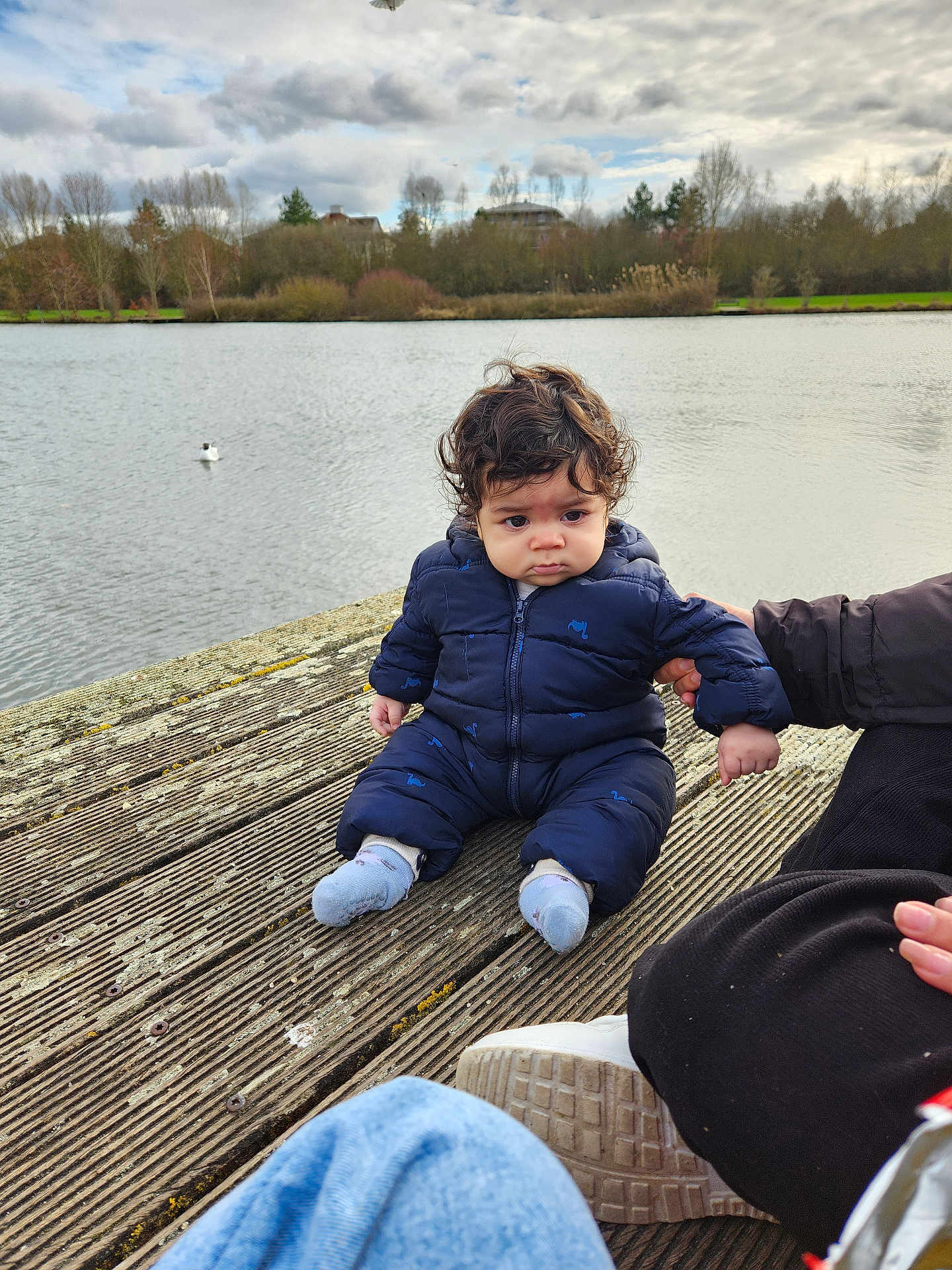 Lucas participe au concours pour gagner de l'argent avec cette photo : baby, toddler, child, dock, wooden_planks, lake, water, jacket, snowsuit, socks, adult_hand, shoe, cloudy_sky, trees, nature, outdoors, portrait, curly_hair, seagull, expression