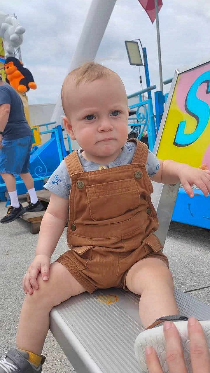 Waylon is registered to the contest to win money with this photo: toddler, child, overalls, bench, amusement_park, outdoor, person, toy, stuffed_animal, blue_shirt, shorts, shoe, hand, sky, metal, sign, adult, casual_clothing, concrete, daytime