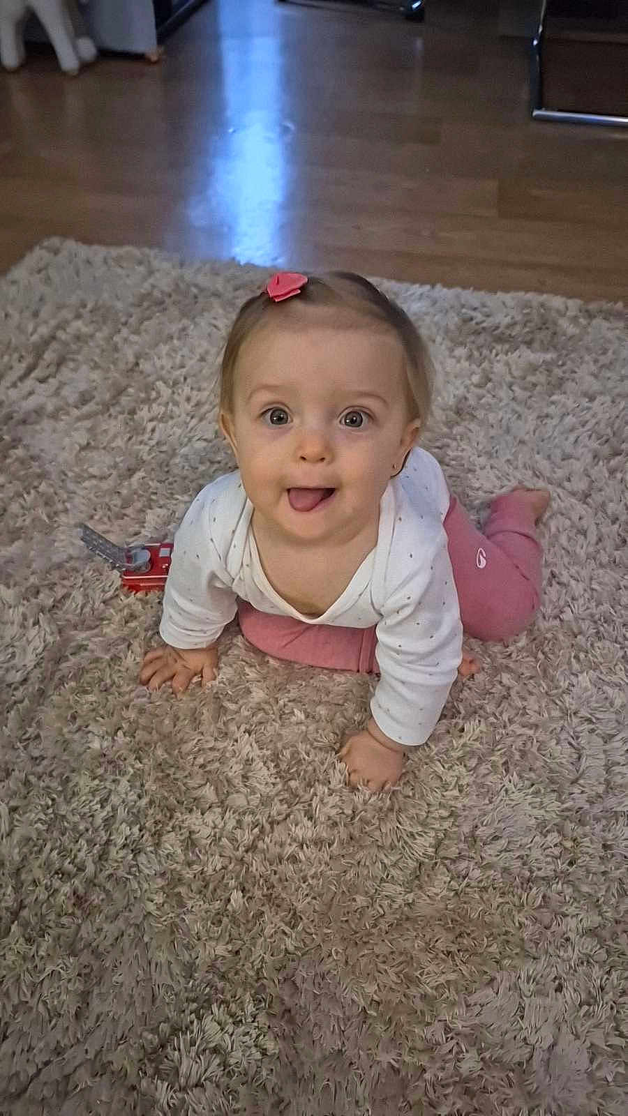 Lyvia a rejoint le concours — aidez-le/la à gagner de superbes lots ! baby, child, crawling, pink_hair_clip, tongue_out, toy, fluffy_rug, beige_rug, wooden_floor, indoor, cute, playful, smiling, person, infant, small_toy, home, floor, clothing, long_sleeve