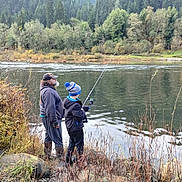 Chris joined the competition — help win amazing prizes! adult, child, fishing_rod, river, water, trees, forest, nature, outdoor, beard, hat, jacket, boots, grass, rocks, overcast, recreation, hobby, family, scenery