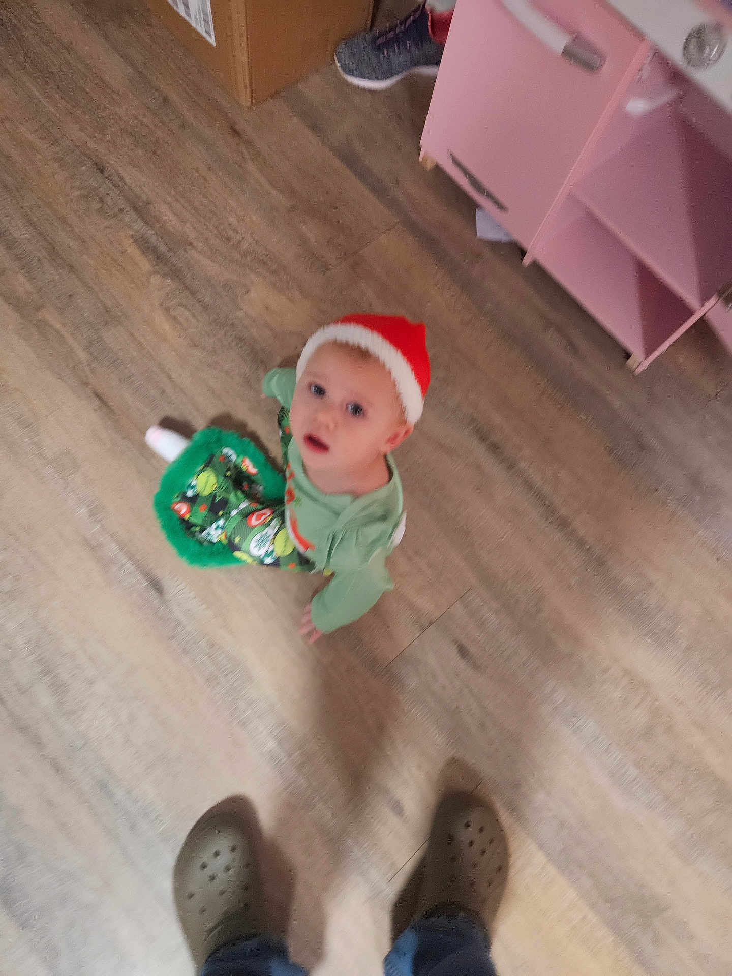 Auttumn is registered to the contest to win money with this photo: child, toddler, floor, wooden_floor, hat, red_hat, green_clothing, festive, indoor, person, footwear, crocs, cabinet, drawer, cardboard_box, toy, curious, looking_up, blur, home