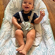 David is registered to the contest to win money with this photo: baby, smiling, child, infant, baby_chair, seatbelt, patterned_fabric, wooden_floor, happy, sitting, indoors, cute, person, face, hand, foot, clothing, home, comfort, expression