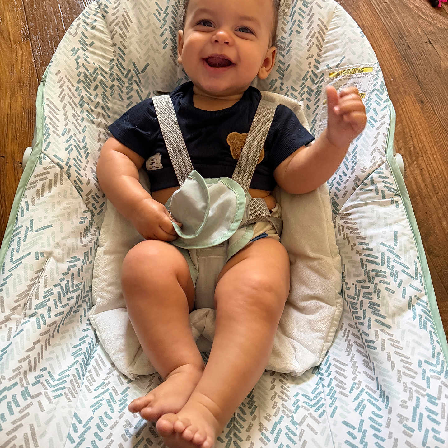 David is registered to the contest to win money with this photo: baby, smiling, child, infant, baby_chair, seatbelt, patterned_fabric, wooden_floor, happy, sitting, indoors, cute, person, face, hand, foot, clothing, home, comfort, expression