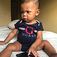 Khyree is registered to the contest to win money with this photo: baby, child, infant, onesie, guess_logo, smartphone, bed, blanket, indoor, curious, expression, person, skin, hair, furniture, hand, sitting, technology, candid, focused