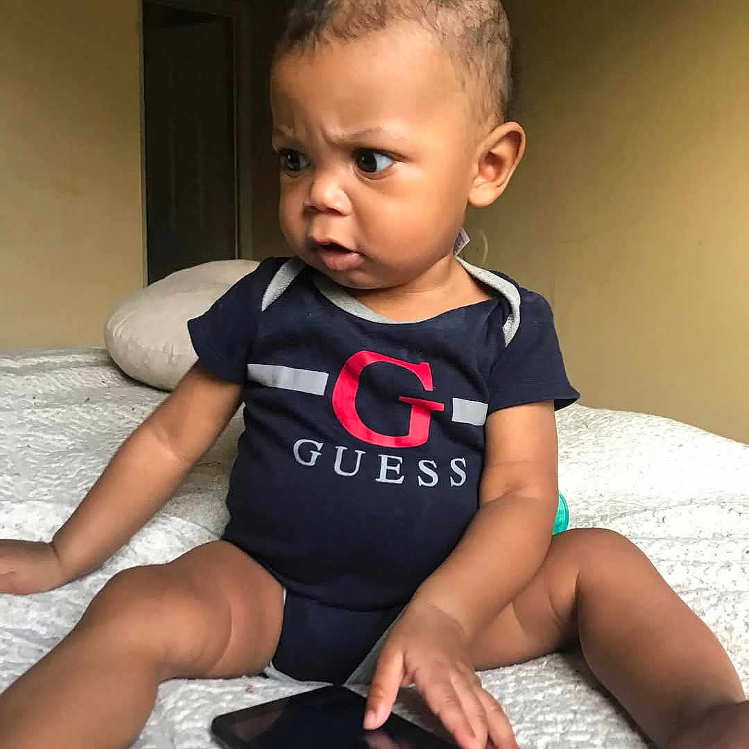 Khyree is registered to the contest to win money with this photo: baby, bed, blanket, candid, child, curious, expression, focused, furniture, guess_logo, hair, hand, indoor, infant, onesie, person, sitting, skin, smartphone, technology