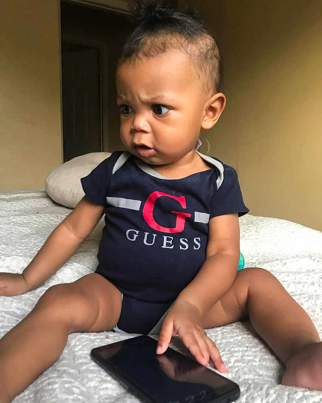 Khyree is registered to the contest to win money with this photo: baby, child, infant, onesie, guess_logo, smartphone, bed, blanket, indoor, curious, expression, person, skin, hair, furniture, hand, sitting, technology, candid, focused