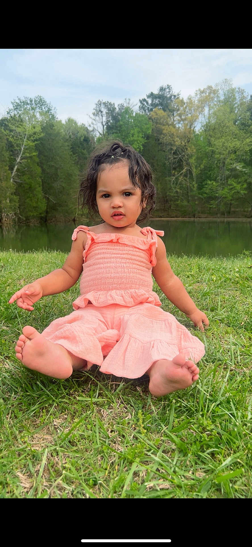 Zaylee joined the competition — help win amazing prizes! toddler, child, grass, lake, water, trees, outdoor, nature, barefoot, curly_hair, pink_clothing, summer, greenery, young_child, portrait, sitting, cute, daylight, park, casual