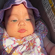 Chloe is registered to the contest to win money with this photo: baby, infant, child, car_seat, purple_hat, floral_dress, sunlight, face, portrait, closeup, cute, soft_skin, warning_label, seatbelt, summer_clothing, outdoor_light, expressionless, headwear, seat, child_safety