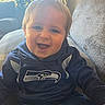 baby, child, infant, smiling, happy, portrait, face, eyes, blond_hair, sitting, couch, home, indoor, lamp, window, sunlight, clothing, shirt, sports_logo, seahawks