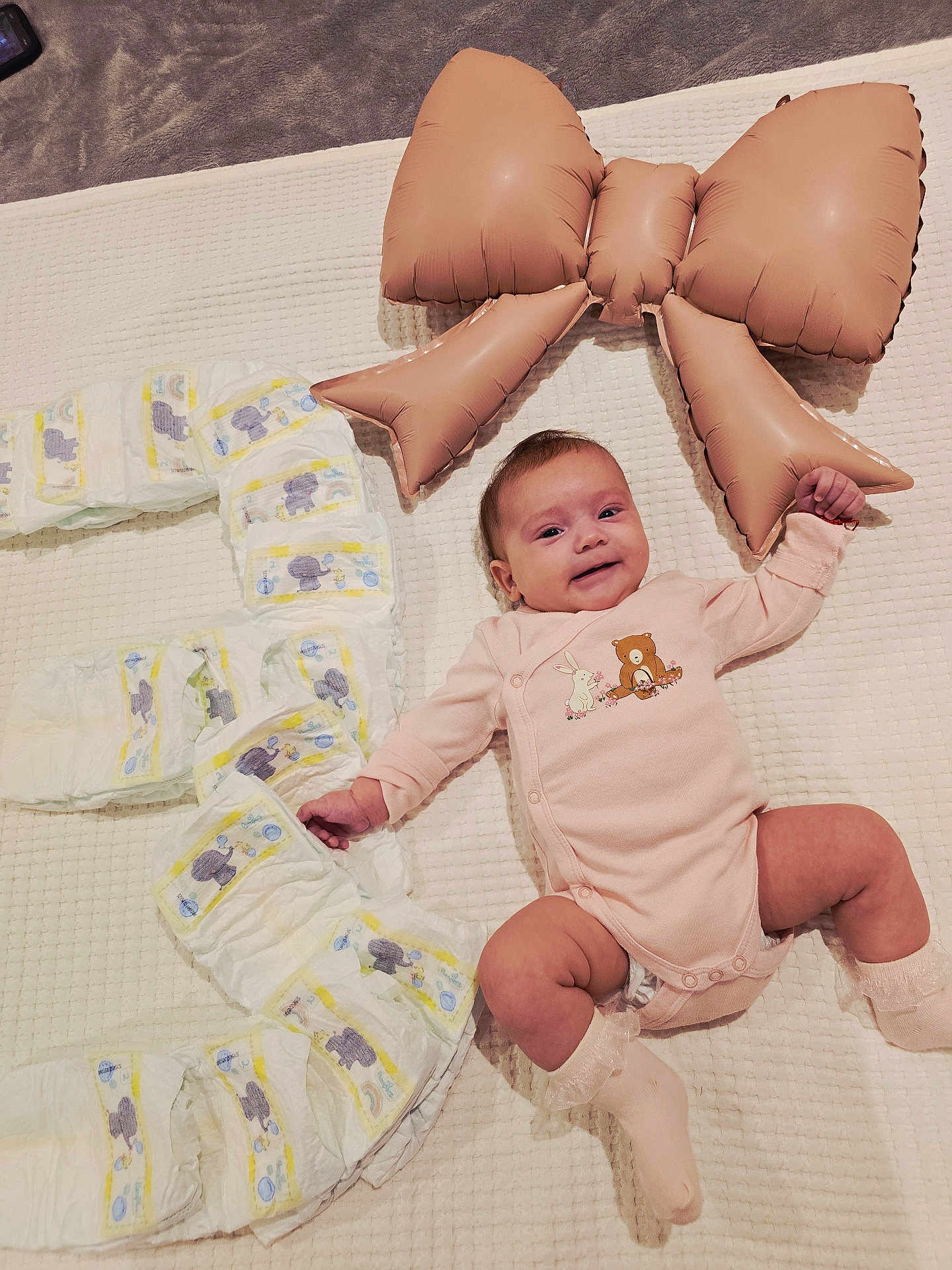 Selena is registered to the contest to win money with this photo: baby, infant, smiling, onesie, pink_clothing, diapers, inflatable_bow, white_blanket, milestone, lying_down, cute, happy, child, baby_sock, indoor, soft_texture, celebration, number_three, baby_face, adorable