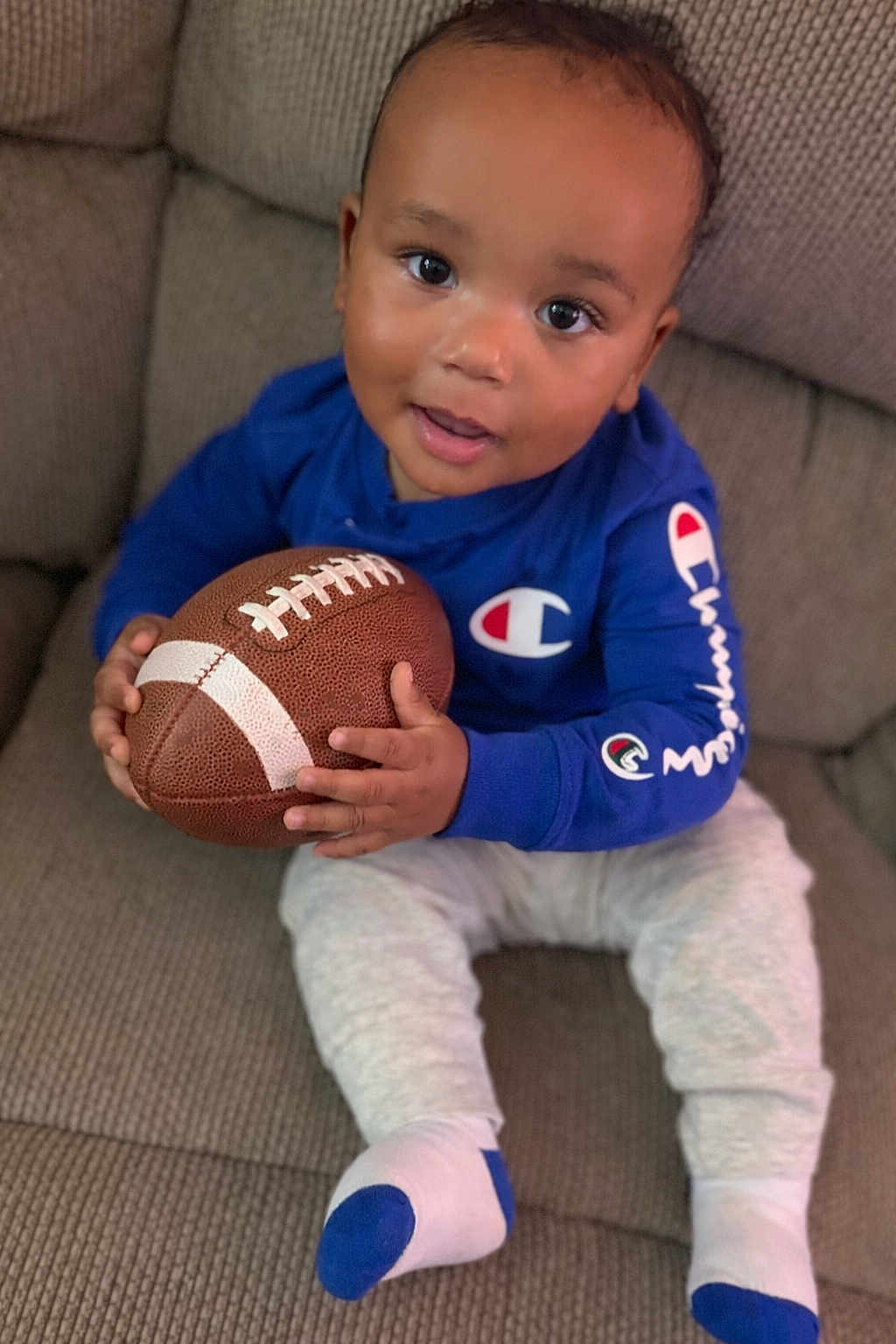 Zyair joined the competition — help win amazing prizes! toddler, child, football, blue_sweatshirt, champion_logo, couch, sitting, white_pants, socks, indoor, cute, baby, smiling, holding_object, face, person, toy, home, playing, happy