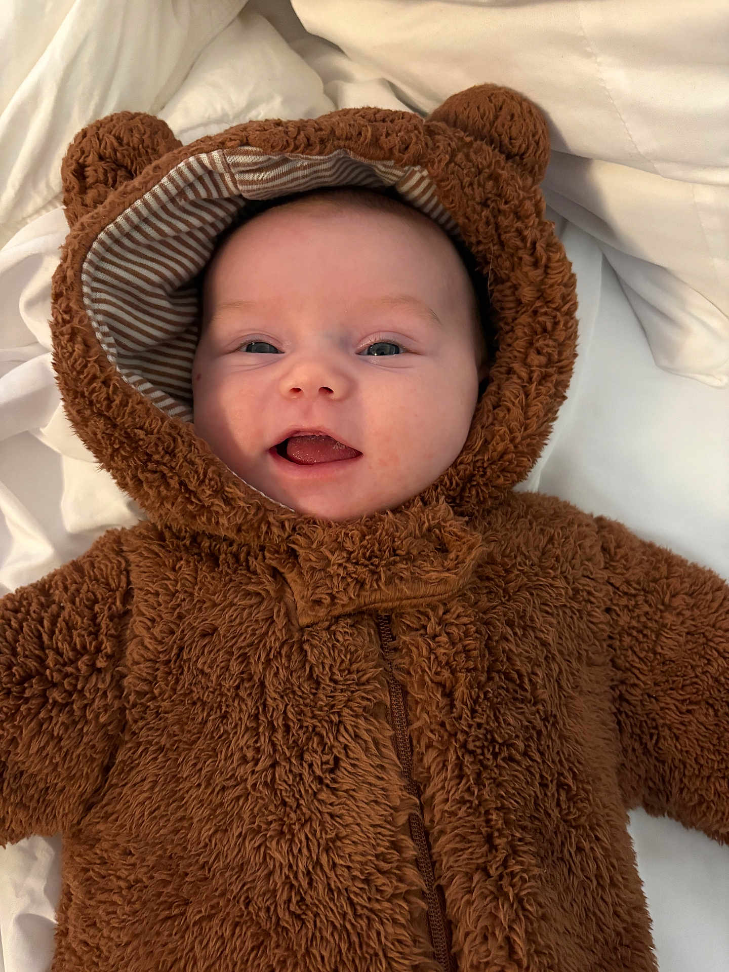 Nellie is registered to the contest to win money with this photo: baby, infant, child, cute, smiling, tongue_out, bear_costume, hood, fuzzy_clothing, soft_texture, white_blanket, portrait, closeup, happy, lying_down, warm_clothing, adorable, face, head, expressive