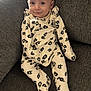 baby, infant, onesie, floral_pattern, couch, fabric, smile, face, person, seated, indoor, cute, child, young, apparel, footed_pajamas, expression, comfort, home, relaxation