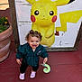 Elyana is registered to the contest to win money with this photo: toddler, child, curly_hair, smiling, pink_shoes, plush_toy, green_jacket, outdoor, red_pavement, pikachu, cartoon, poster, plant_pot, plant, happy, person, casual_clothing, daylight, cute, playful