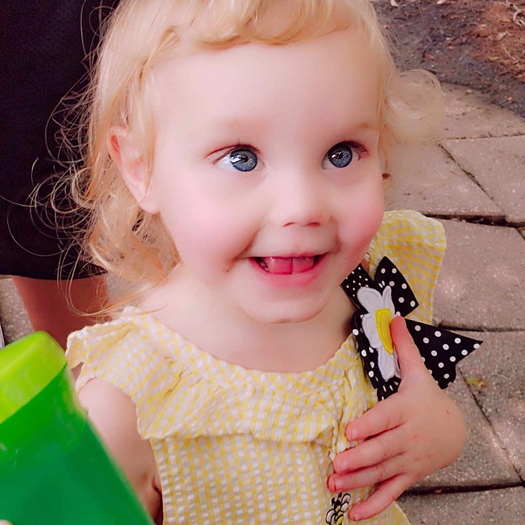 Celestia joined the competition — help win amazing prizes! bee_embroidery, blonde_hair, blue_eyes, child, clothing, curly_hair, daisy, green_cup, hand, happy, leaves, outdoor, pavement, person, polka_dot_bow, portrait, smiling, soil, toddler, yellow_dress