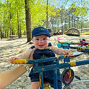 Maxwell joined the competition — help win amazing prizes! baby, child, denim_overalls, cap, playground, tricycle, sand, trees, greenery, sunlight, hand, outdoor, park, smile, happy, person, toy, blue, yellow, nature