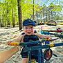 baby, child, denim_overalls, cap, playground, tricycle, sand, trees, greenery, sunlight, hand, outdoor, park, smile, happy, person, toy, blue, yellow, nature