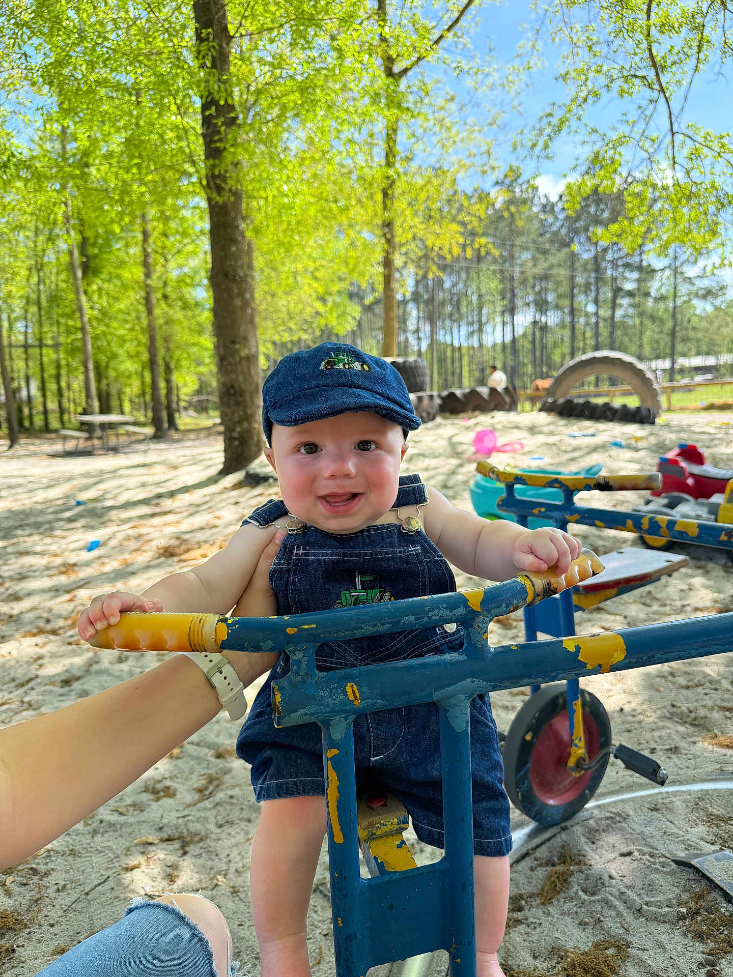 Maxwell joined the competition — help win amazing prizes! baby, child, denim_overalls, cap, playground, tricycle, sand, trees, greenery, sunlight, hand, outdoor, park, smile, happy, person, toy, blue, yellow, nature
