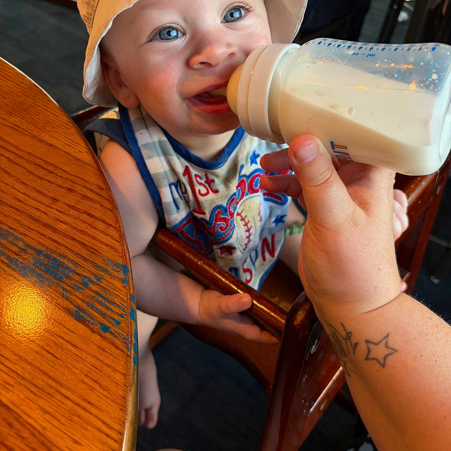 Grayson is registered to the contest to win money with this photo: baby, bottle, milk, feeding, high_chair, child, hat, bucket_hat, tattoo, hand, table, wood, blue_eyes, smiling, indoor, clothing, person, infant, cute, dining