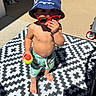 toddler, child, hat, bucket_hat, sunglasses, red_sunglasses, tennis_ball, barefoot, shorts, patterned_rug, outdoor, patio, sunny, shadow, concrete, door, hose, summer, playful, person