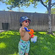 Zayden is registered to the contest to win money with this photo: toddler, child, grass, outdoor, sunlight, hat, sunglasses, toy, water_gun, shorts, tree, fence, summer, play, greenery, barefoot, person, casual, daylight, nature