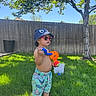toddler, child, grass, outdoor, sunlight, hat, sunglasses, toy, water_gun, shorts, tree, fence, summer, play, greenery, barefoot, person, casual, daylight, nature