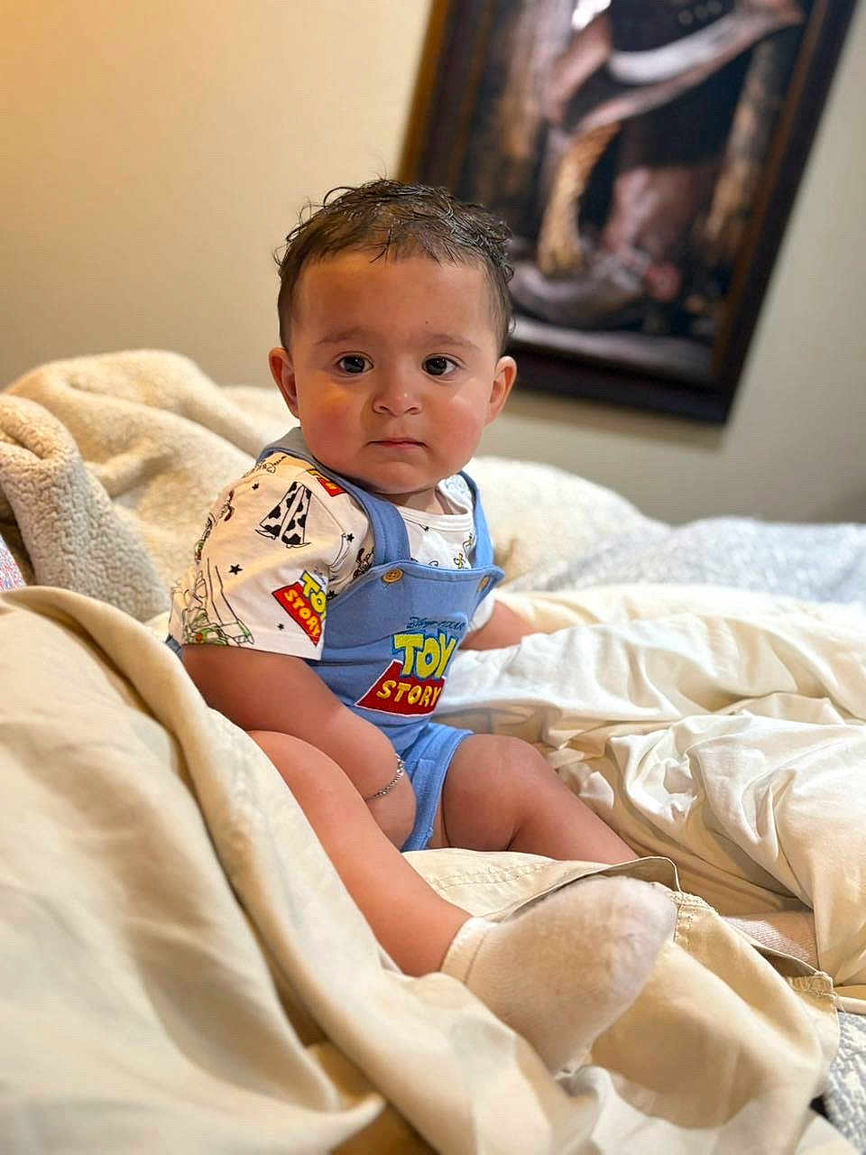 David is registered to the contest to win money with this photo: baby, child, toddler, face, clothing, toy_story, overalls, blanket, bed, indoor, portrait, sitting, cute, expression, person, young_child, soft, comfort, casual, curious