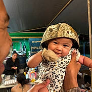 Suliana is registered to the contest to win money with this photo: baby, smiling, camo_hat, onesie, adult_hands, outdoor, night, tent, people, gathering, table, food, drinks, casual, portrait, happy, cute, infant, holding, celebration