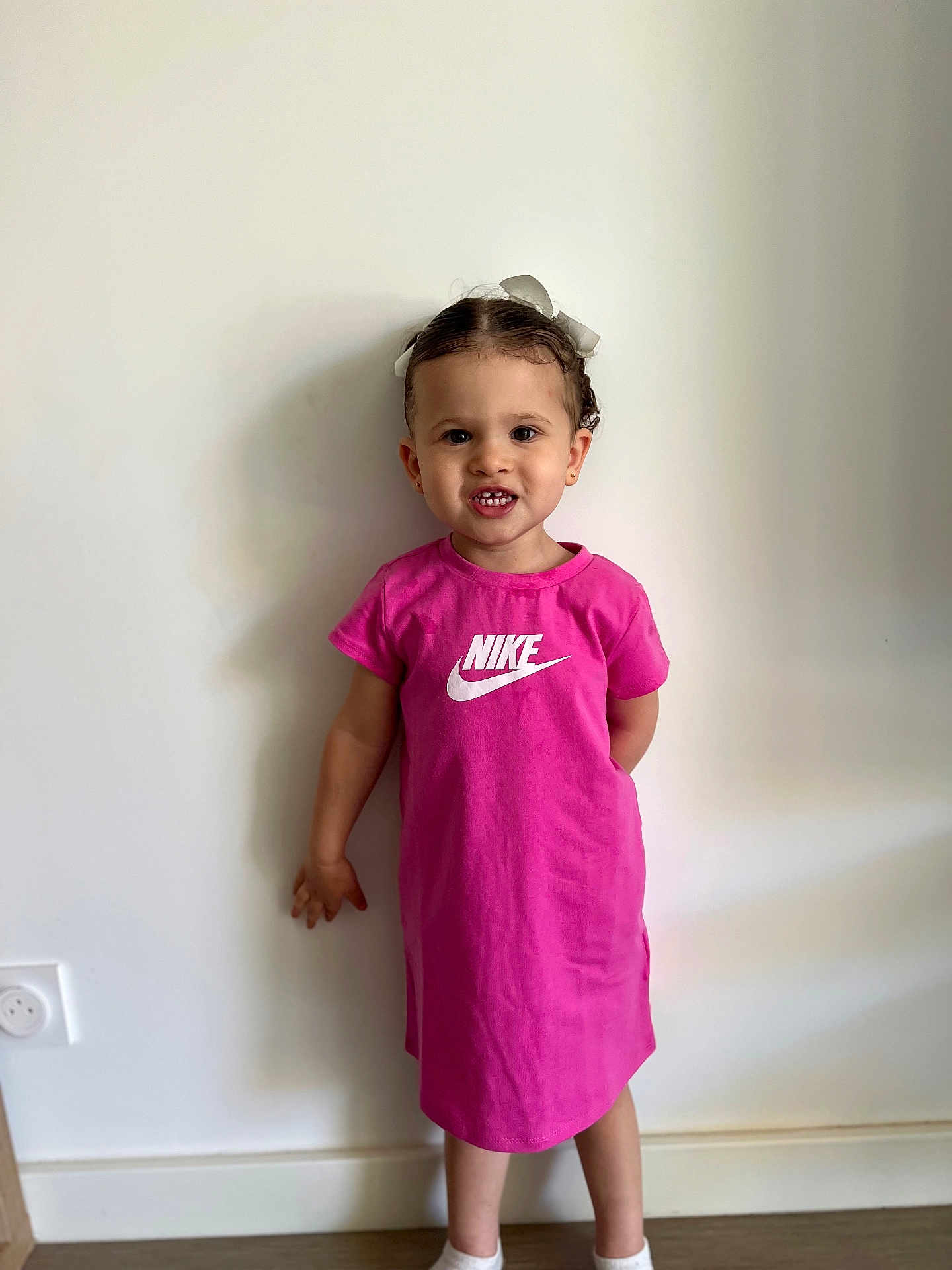 Kataleya a rejoint le concours — aidez-le/la à gagner de superbes lots ! toddler, child, girl, pink_dress, nike_logo, white_bow, standing, indoor, smile, teeth, hair, face, wall, floor, socks, wooden_floor, earrings, shadow, casual_clothing, portrait