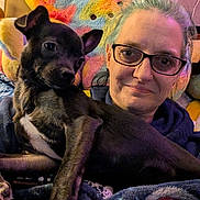 Luna is registered to the contest to win money with this photo: person, dog, glasses, indoor, blanket, plush, couch, smile, pet, animal, colorful, pillow, furniture, snuggle, cozy, brown_hair, black_dog, casual, relaxation, home