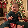 baby, child, christmas_tree, gift, pajamas, holiday, festive, decorations, stockings, fireplace, wrapped_presents, indoors, smiling, feet, face, sitting, holiday_lights, red, green, cozy