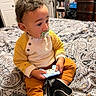 toddler, child, baby, portrait, sitting, pacifier, toy_controller, gaming_toy, bedroom, bed, paisley_bedding, yellow_clothing, orange_pants, black_socks, curly_hair, indoors, bookshelf, dresser, curious, family_photo