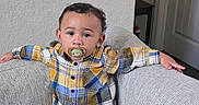 Elias is registered to the contest to win money with this photo: child, toddler, baby, standing, couch, sofa, indoors, portrait, pacifier, plaid_shirt, white_pants, socks, arms_outstretched, serious_expression, face, hair, furniture, living_room, door, wall