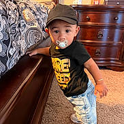 Elias is registered to the contest to win money with this photo: child, toddler, baby, portrait, standing, pacifier, baseball_cap, t_shirt, jeans, sneakers, bracelet, bedroom, bed, dresser, furniture, carpet, indoors, patterned_bedding, looking_at_camera, home