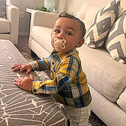 Elias is registered to the contest to win money with this photo: baby, child, toddler, standing, pacifier, smiling, portrait, indoors, living_room, sofa, couch, ottoman, cushions, plaid_shirt, pants, home, furniture, carpet, lamp, daylight