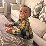 baby, child, toddler, standing, pacifier, smiling, portrait, indoors, living_room, sofa, couch, ottoman, cushions, plaid_shirt, pants, home, furniture, carpet, lamp, daylight
