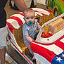 baby, child, pacifier, kiddie_ride, toy_car, usa_flag, indoor, mall, adult, shorts, sneakers, floor, hand, seat, steering_wheel, striped_shirt, expression, footwear, person, playful
