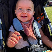 Dalton joined the competition — help win amazing prizes! baby, smile, teeth, stroller, outdoor, sunlight, child, happy, infant, seatbelt, clothing, pink_bib, gray_shirt, hand, grass, toy, face, person, portrait, daytime