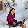 child, smiling, sled, snow, winter, boots, long_hair, outdoor, holiday, joy, tree, lantern, blurred_background, cold_weather, casual_clothing, sitting, fun, playful, snowy_scene, happy