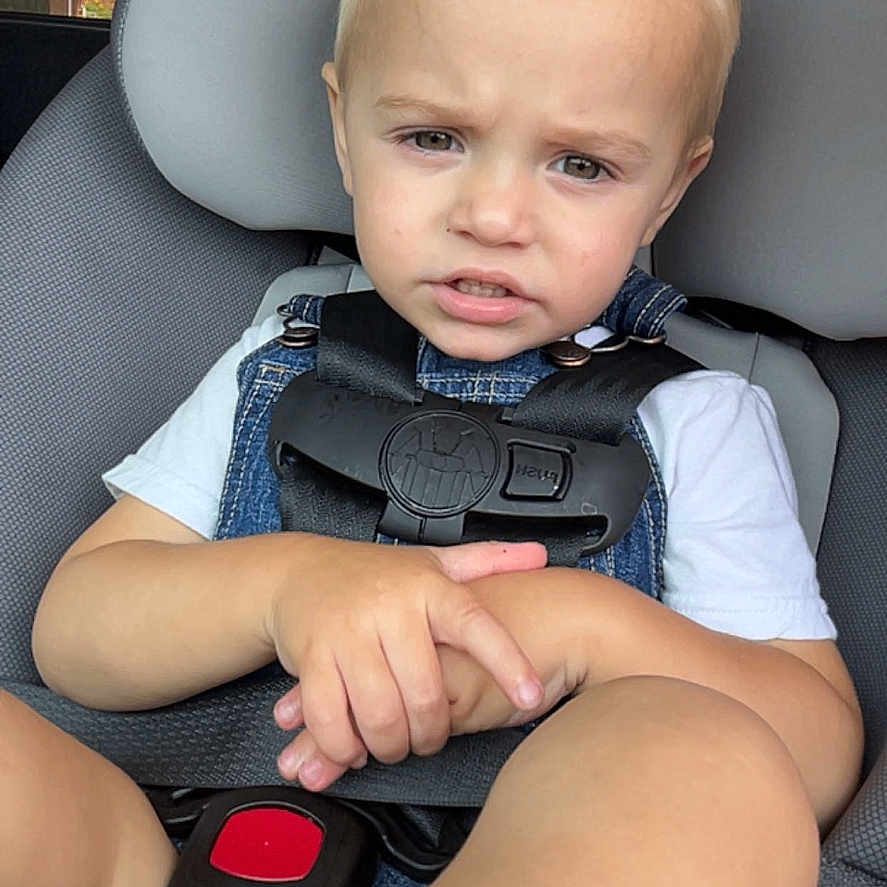 Pave is registered to the contest to win money with this photo: blonde_hair, car, car_seat, child, daylight, denim_overalls, face, hands, person, road, safety_harness, seat, seatbelt, serious_expression, toddler, transportation, vehicle_interior, white_shirt, window, young_child