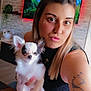 woman, dog, chihuahua, tattoo, living_room, television, table, plant, shelf, black_top, long_hair, portrait, pet, indoor, smiling, cute, small_dog, holding, face, casual